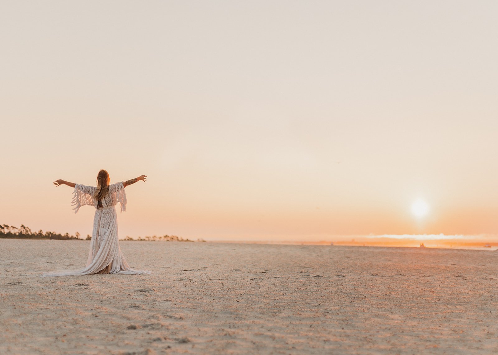 PCB Photographers collab. Girl stands wearing white dress, arms outstretched facing rising sun in st. andrews state park.