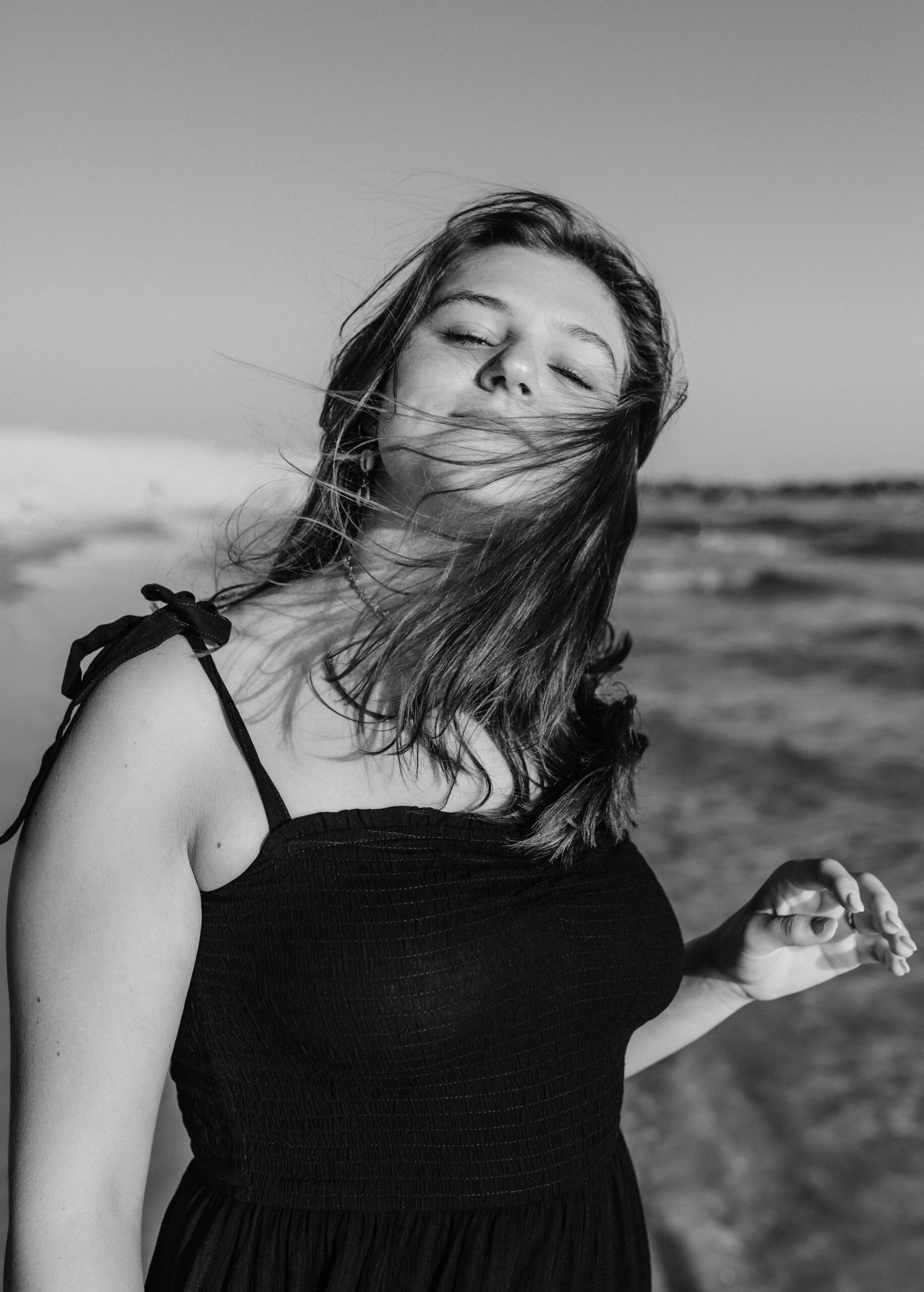 senior girl posing in black dress on beach - with wind blowing hair back. Senior session by Brittney Stanley in St. Andrews State Park
