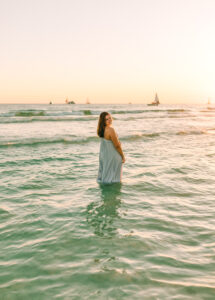 highschool senior portraits of brunette girl - in grey dress, standing in teal colored waters with boats in the distant background at sunset on PCB Florida