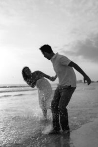 Newly engaged young couple hold hands & play on beach in PCB Florida - B&W edit of image taken by photographer Brittney Stanley of Be Seen Photos