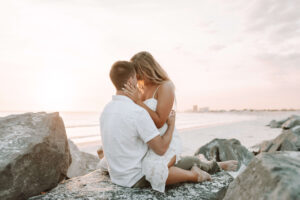 Newly engaged young couple cuddle & kiss intimately on rocks at sunset on beach in PCB Florida