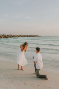 Young man kneels & proposes to girlfriend on edge of beach/water in PCB Florida