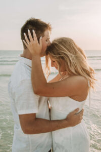 newly engaged young couple wearing white & tan embrace & kiss beachside in PCB Florida