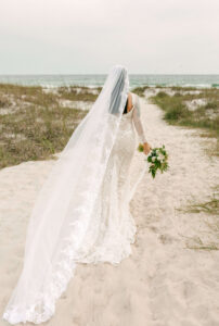 Bride walking down sand path in West PCB, Florida - wearing gown & holding flowers